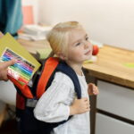 Un parent glisse des feutres de couleur et un cahier dans le sac à dos d'une petite fille blonde prête pour la rentrée, illustrant comment choisir le bon sac d’école pour son enfant en maternelle?