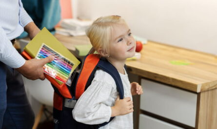 Un parent glisse des feutres de couleur et un cahier dans le sac à dos d'une petite fille blonde prête pour la rentrée, illustrant comment choisir le bon sac d’école pour son enfant en maternelle?