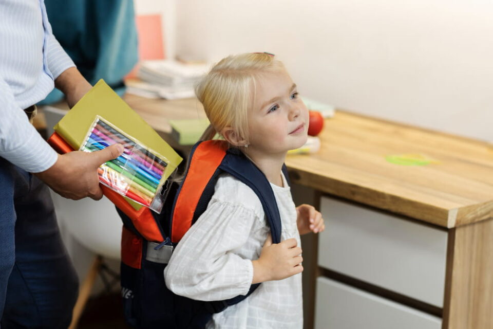 Un parent glisse des feutres de couleur et un cahier dans le sac à dos d'une petite fille blonde prête pour la rentrée, illustrant comment choisir le bon sac d’école pour son enfant en maternelle?